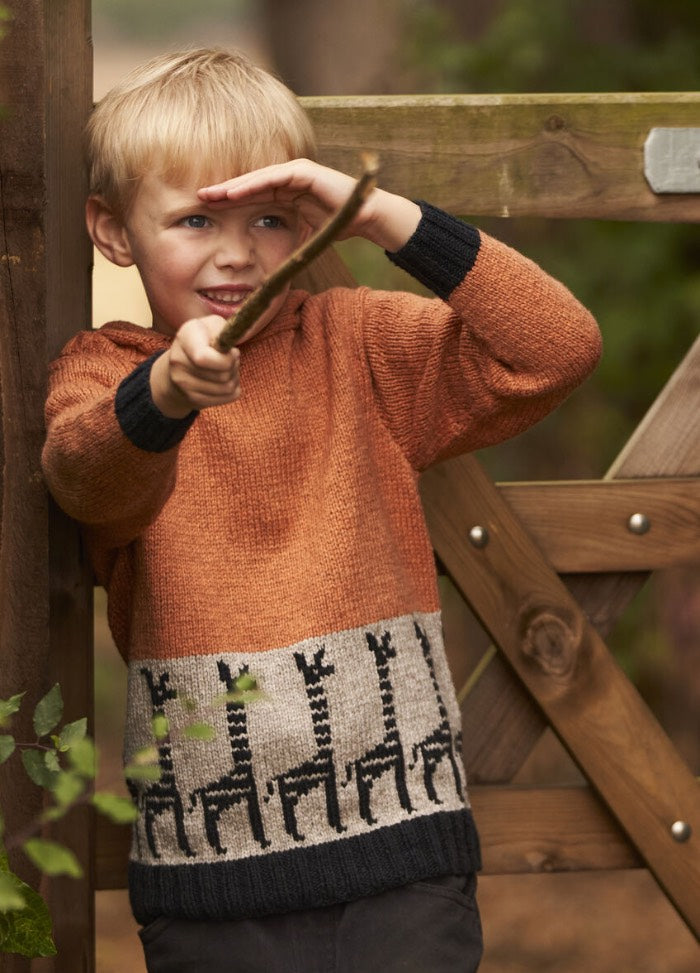 A child with blond hair wears an orange and gray sweater with giraffe patterns, playfully holding a stick like a telescope, smiling near a wooden gate.