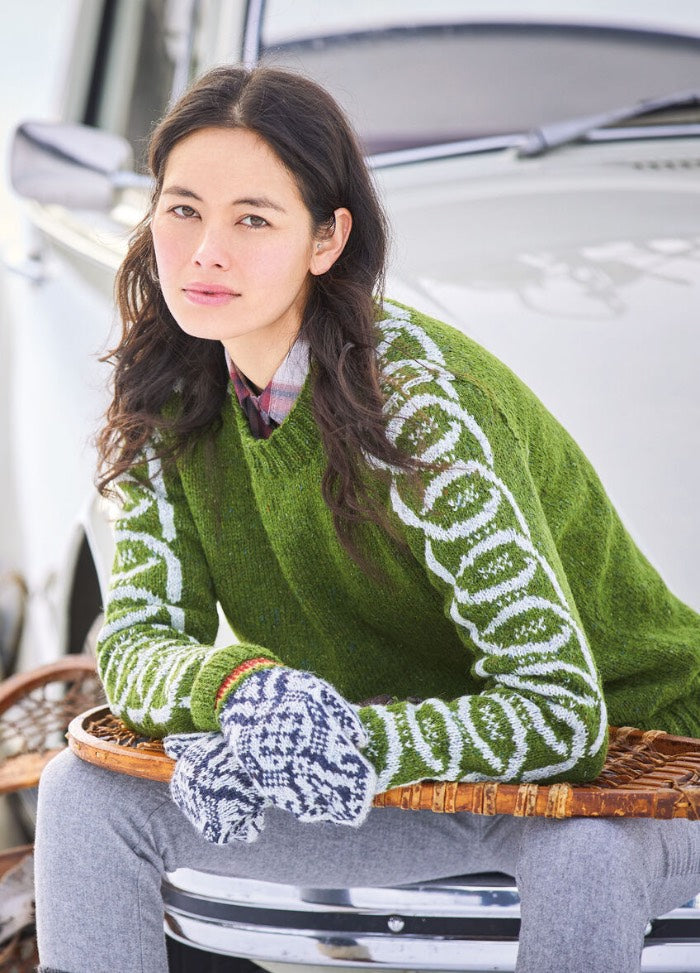 Young woman in a green sweater with patterned sleeves leans against a car, wearing gray mittens and gray pants, exuding a relaxed and thoughtful vibe.