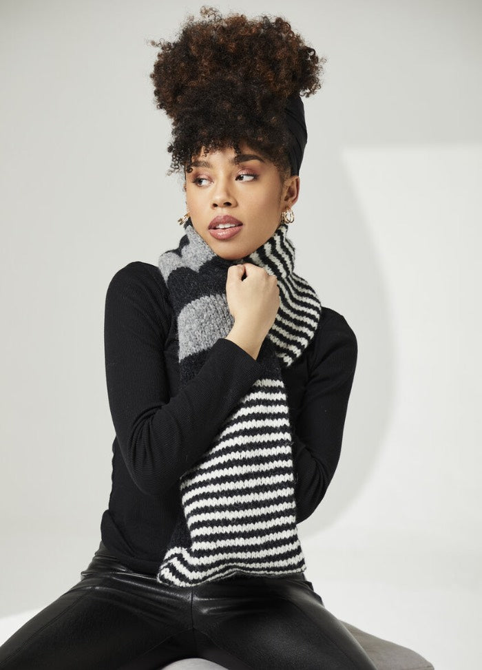 A woman with curly hair wears a black top and leather pants, wrapped in a striped black-and-white scarf, gazing to the side in a neutral setting.