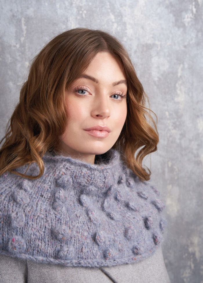 Woman with wavy brown hair wearing a textured, light blue knitted shawl over a grey sweater. She looks calm with a neutral expression.
