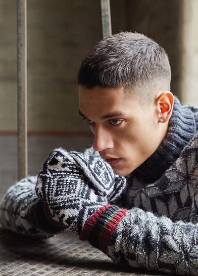 A person in a patterned sweater and gloves rests their head thoughtfully on their hands. The muted background and soft light create a contemplative mood.