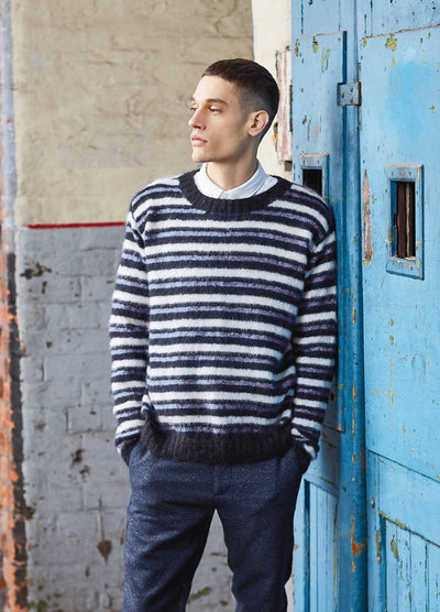 A person in a striped sweater and collared shirt stands by a weathered blue door, looking thoughtfully to the side in an industrial setting.