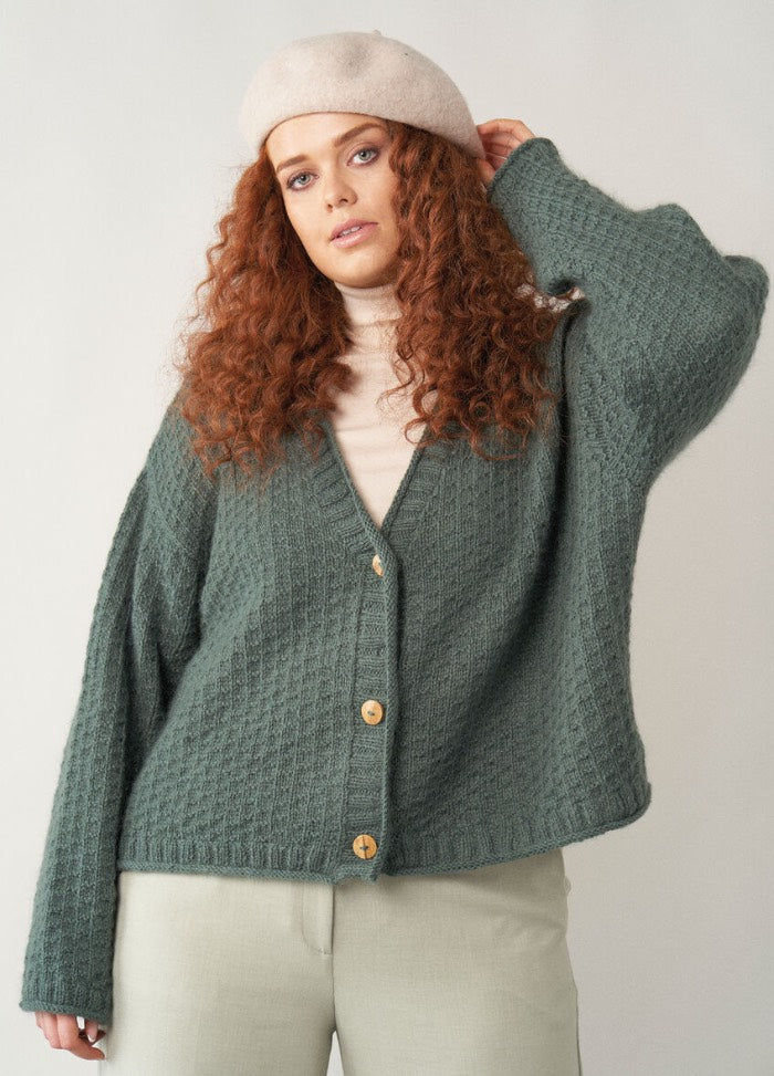A relaxed crew-neck cardigan in muted pink, featuring wide ribbed cuffs and hem, styled with rolled-up sleeves for a laid-back look.