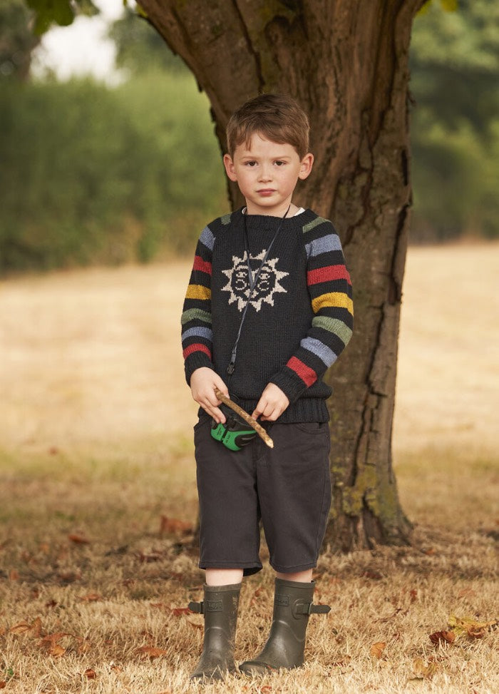 A young boy stands under a tree on dry grass. He's wearing a sweater with a sun design and rainbow-striped sleeves, dark shorts, and boots, exuding a contemplative mood.