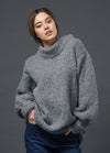 Woman in a cozy gray turtleneck sweater and blue jeans poses against a plain dark gray background, conveying a relaxed, casual vibe.