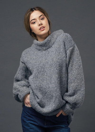 Woman in a cozy gray turtleneck sweater and blue jeans poses against a plain dark gray background, conveying a relaxed, casual vibe.