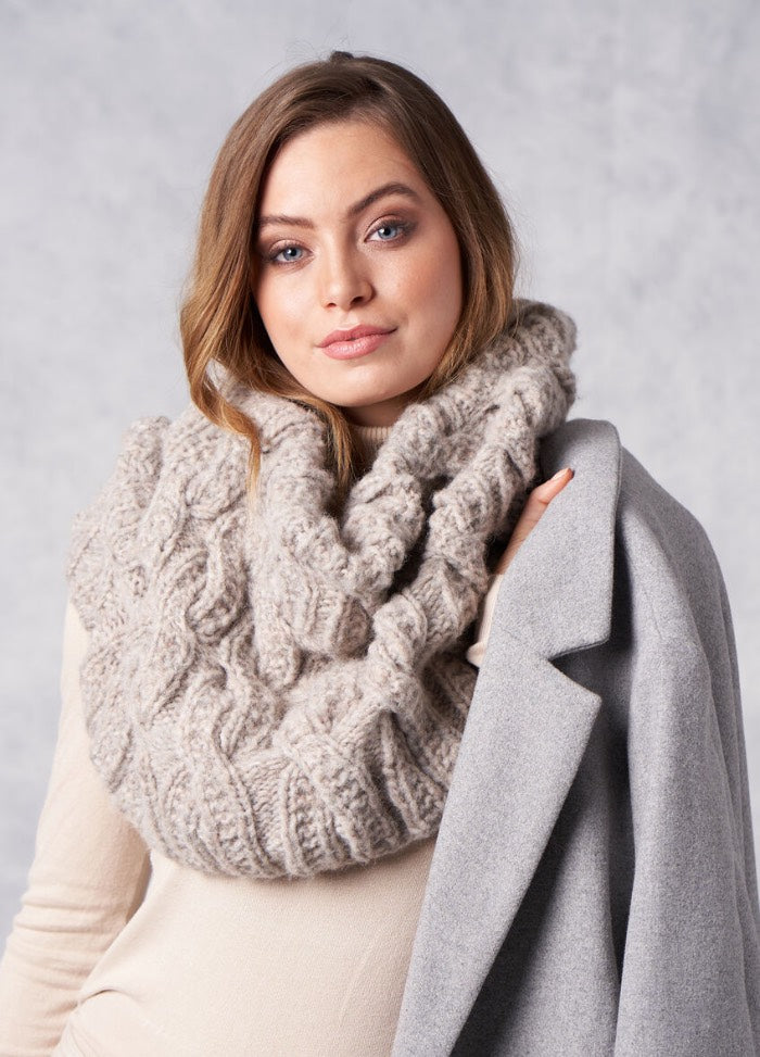 A woman with long brown hair wearing a cozy, chunky knit scarf and holding a draped gray coat over her shoulder, set against a neutral background.