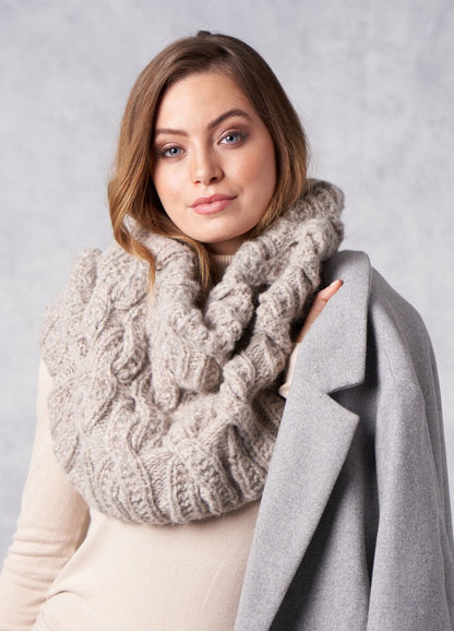 A woman with long brown hair wearing a cozy, chunky knit scarf and holding a draped gray coat over her shoulder, set against a neutral background.
