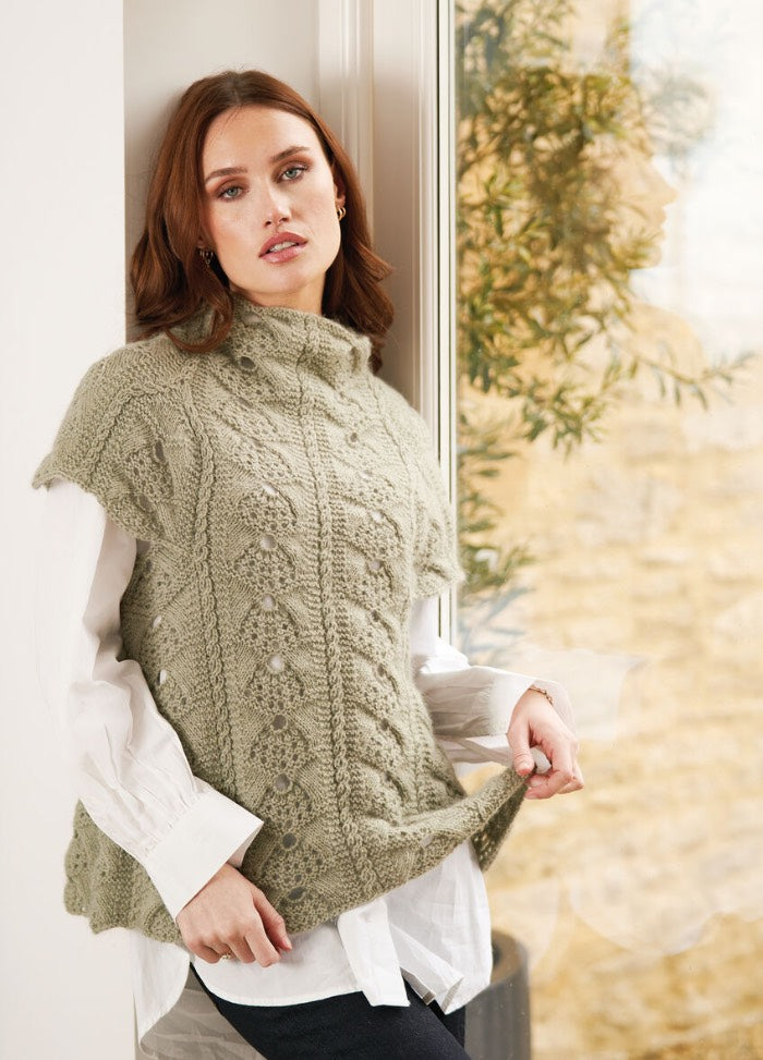 Woman with medium-length hair stands by a window, wearing a textured, green knitted vest over a white shirt. She looks relaxed and thoughtful.