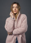 Young woman with long hair wearing a cozy, pink, chunky knit sweater poses confidently with a thoughtful expression against a dark gray background.