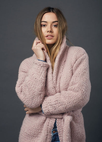 Young woman with long hair wearing a cozy, pink, chunky knit sweater poses confidently with a thoughtful expression against a dark gray background.