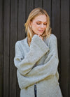 Woman in a cozy gray sweater stands against a dark wooden wall, looking down with a gentle smile. The mood is serene and reflective.