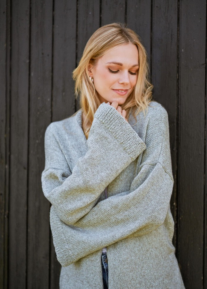 Woman in a cozy gray sweater stands against a dark wooden wall, looking down with a gentle smile. The mood is serene and reflective.