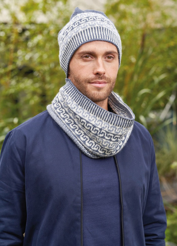 A man wearing a cozy, patterned knit hat and a matching cowl, both in shades of blue and gray, standing outdoors.