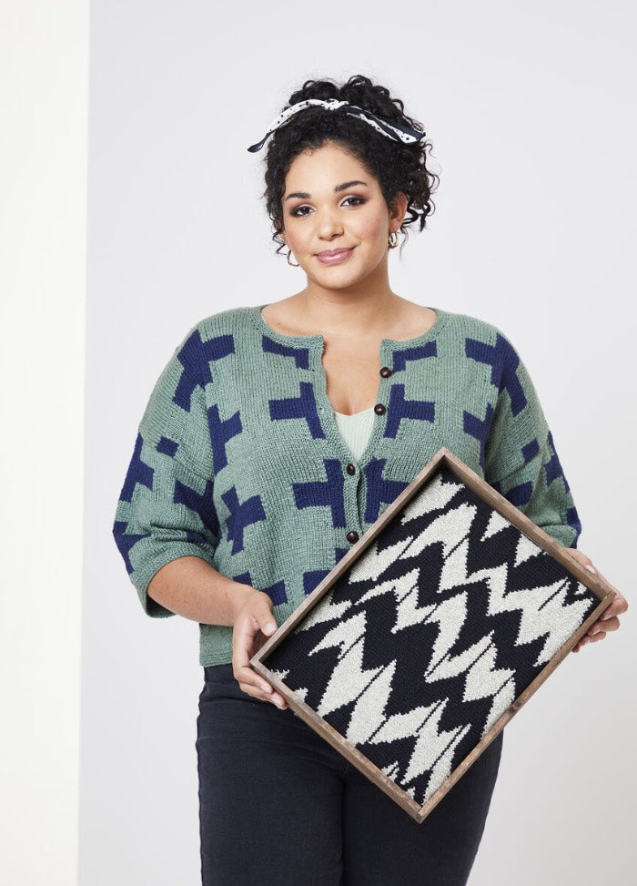 A woman stands holding a framed black and white zigzag patterned item. She wears a green and navy geometric sweater, smiling in a neutral setting.