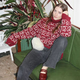 A woman in an oversized Heart Together Sweater sits on a green couch, showcasing its boxy fit and wide sleeves.