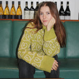 A woman sits comfortably on a green couch, showcasing the oversized Heart Together Sweater with a big turtleneck.