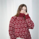 A woman showcases a boxy red sweater with heart patterns, designed with wide sleeves and a cozy turtleneck.