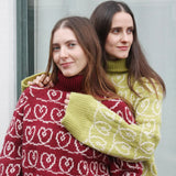 Two women wearing oversized sweaters adorned with hearts, one in green and the other in red, featuring wide sleeves and turtlenecks.