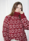 A woman with long brown hair is wearing a red turtleneck sweater with white intertwined heart patterns. She gently holds the collar, exuding a cozy, warm vibe.