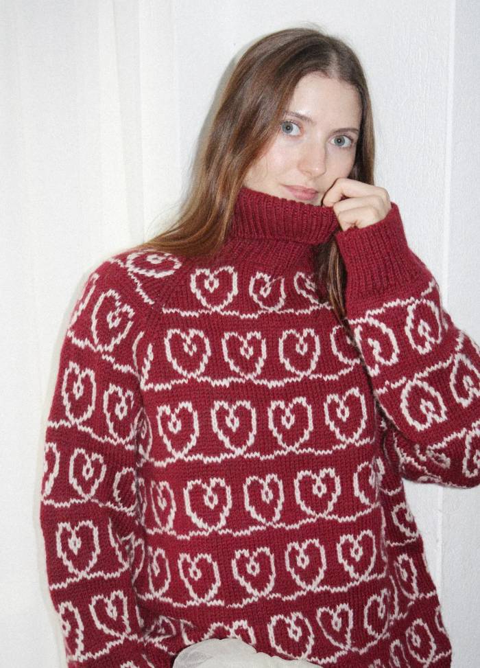 A woman with long brown hair is wearing a red turtleneck sweater with white intertwined heart patterns. She gently holds the collar, exuding a cozy, warm vibe.