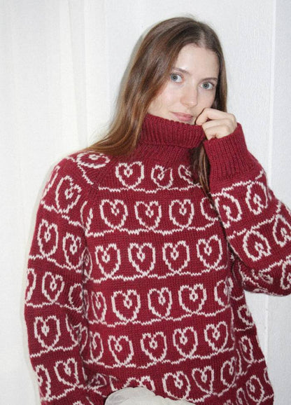 A woman with long brown hair is wearing a red turtleneck sweater with white intertwined heart patterns. She gently holds the collar, exuding a cozy, warm vibe.