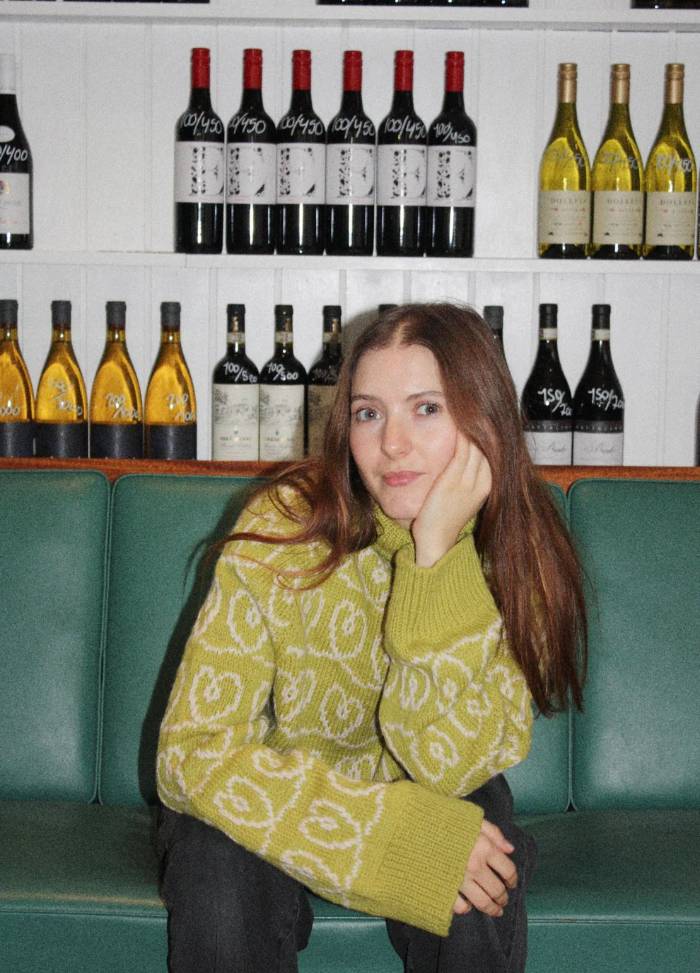 A young woman with long hair sits on a green bench, wearing a yellow patterned sweater, her chin resting on her hand. Behind her are shelves of wine bottles, creating a relaxed and cozy atmosphere.