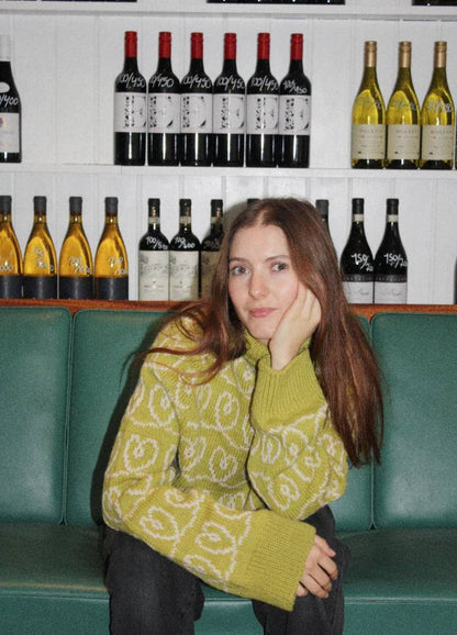 A young woman with long hair sits on a green bench, wearing a yellow patterned sweater, her chin resting on her hand. Behind her are shelves of wine bottles, creating a relaxed and cozy atmosphere.