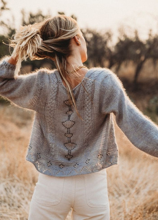 A woman stands in a field, facing away. She's wearing a knitted sweater with intricate back detailing and white pants. Her hair is tied up, and the setting sun casts a warm glow, evoking a serene, peaceful mood.
