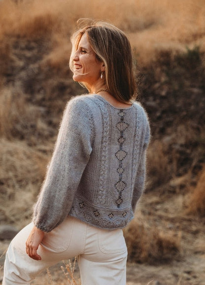 Woman smiling in a knit sweater with intricate patterns, standing outdoors in a dry, grassy field. The atmosphere is warm and serene.