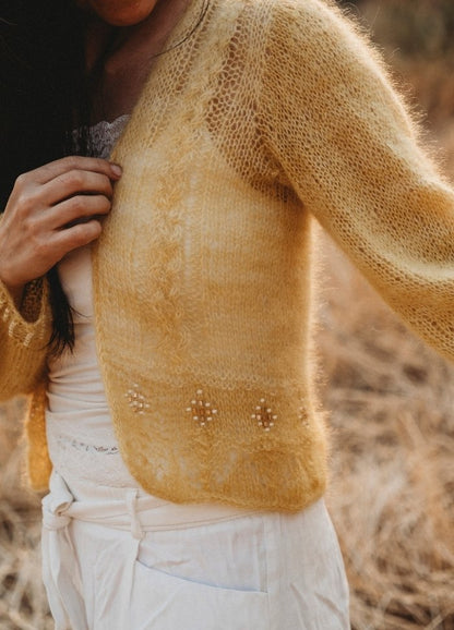 A person in a cozy yellow knit sweater with delicate embroidery holds the front. The background features soft, blurred golden grass, evoking a warm, autumnal feel.