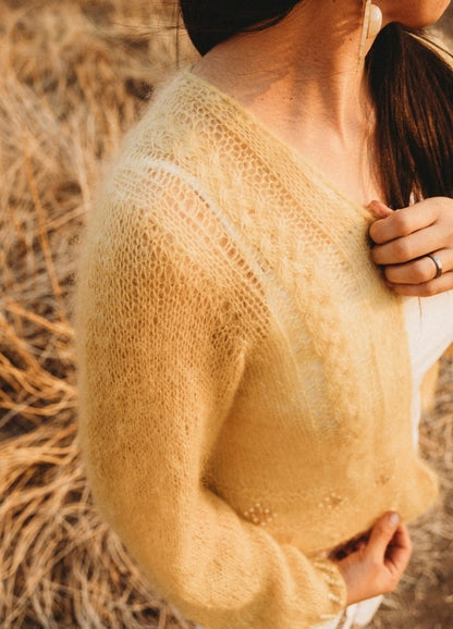 A person in a cozy, light yellow knit sweater stands in a dry, grassy area. The scene is warm and serene, with soft lighting and autumn vibes.