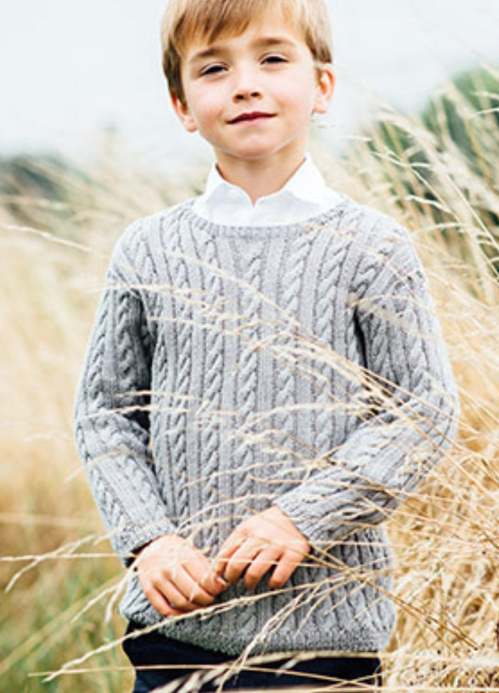 Henry Sweater: Person standing outdoors in tall grass, wearing a grey hand-knitted cable-knit sweater over a white collared shirt.
