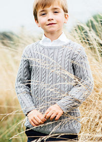 Henry Sweater: Person standing outdoors in tall grass, wearing a grey hand-knitted cable-knit sweater over a white collared shirt.