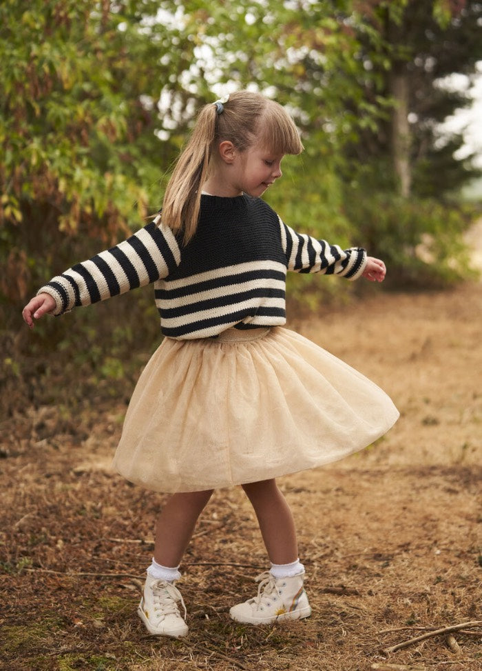 A young girl twirls joyfully on a dirt path, wearing a striped sweater and a beige tulle skirt, surrounded by greenery. Her playful expression and pose convey happiness.