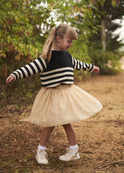 A young girl twirls joyfully on a dirt path, wearing a striped sweater and a beige tulle skirt, surrounded by greenery. Her playful expression and pose convey happiness.