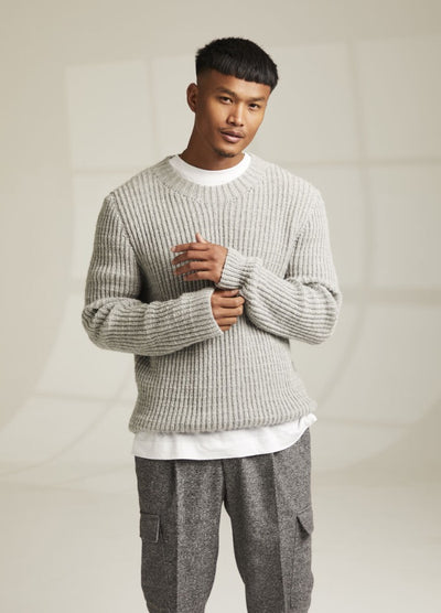Young man in a light grey ribbed sweater over a white shirt, and grey cargo pants. He stands in a softly lit room with a neutral background, looking relaxed.