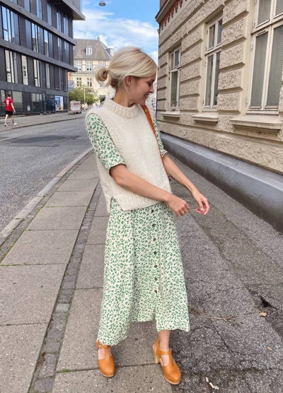 Woman wearing a light green floral dress, cream knit vest, and tan shoes stands on a city sidewalk. She is smiling and looking down, exuding a playful, casual vibe.