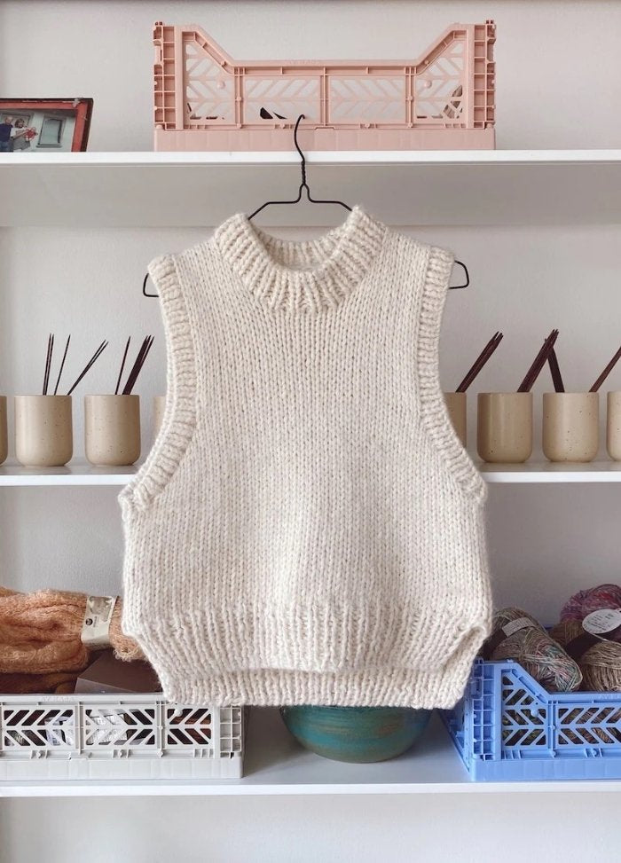 A cream knit sleeveless sweater vest hangs on a hanger against a white shelf. 