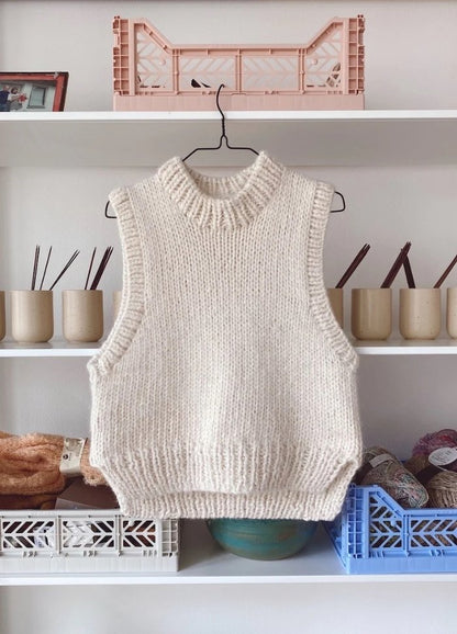 A cream knit sleeveless sweater vest hangs on a hanger against a white shelf. 