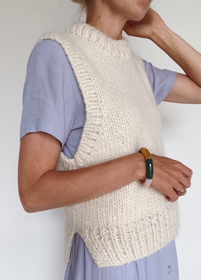 A person in a light purple dress wears a chunky, white knitted vest. Their hand rests on their neck, revealing colorful bangles. The tone is cozy and stylish.