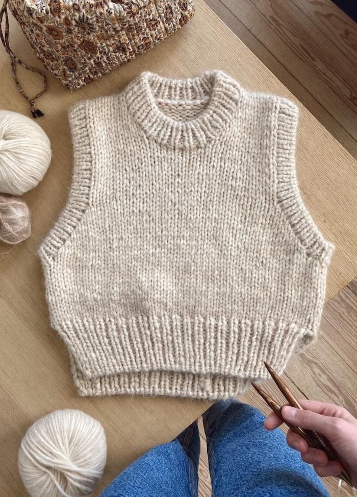 Beige hand-knitted sleeveless sweater on a wooden table, surrounded by yarn balls and knitting needles. Casual, cozy crafting vibe.