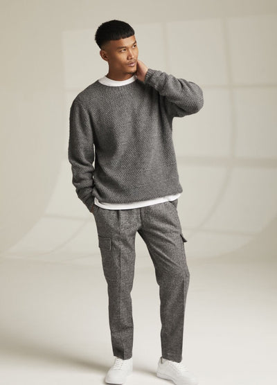 Man wearing a gray textured sweater, white T-shirt, gray cargo pants, and white sneakers in a bright, minimalist setting, exuding a casual tone.