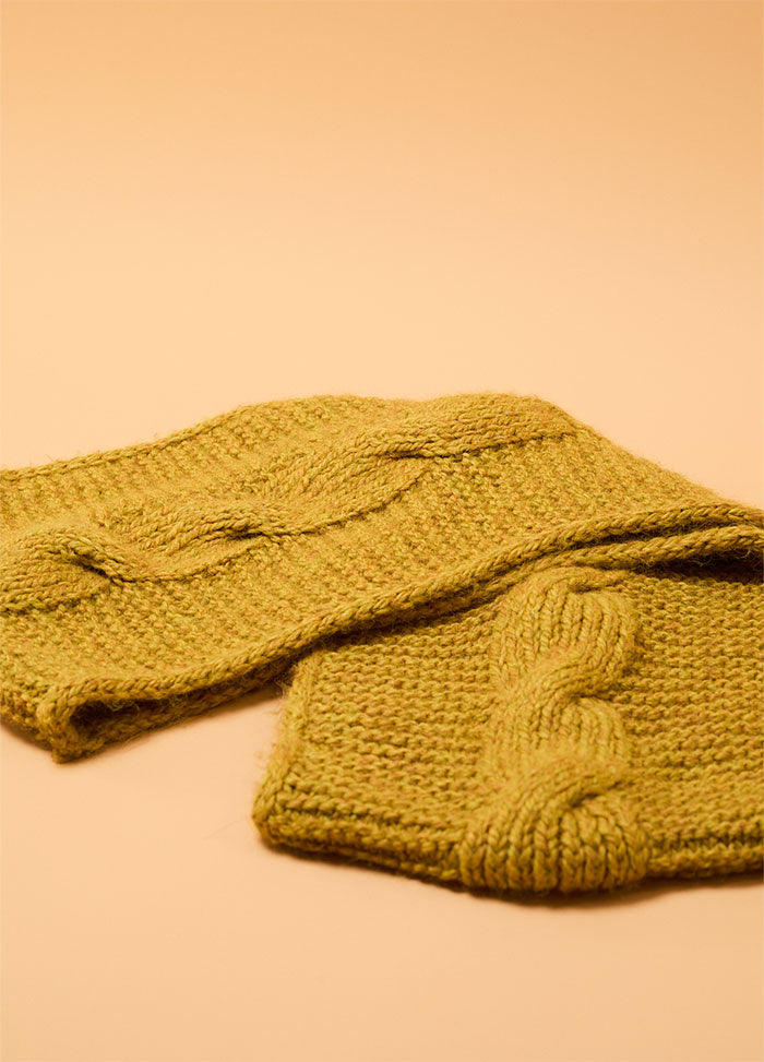 Folded Honey Cable Cowl in mustard yellow yarn, showing wide braided cable stitches against a textured knit background, displayed on a warm beige surface.