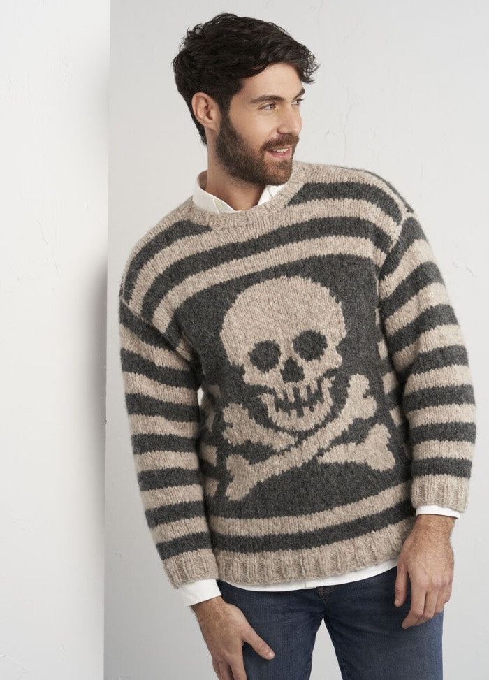 Man in a cozy, striped sweater with a skull and crossbones design leans casually against a white wall, exuding a relaxed, stylish vibe.