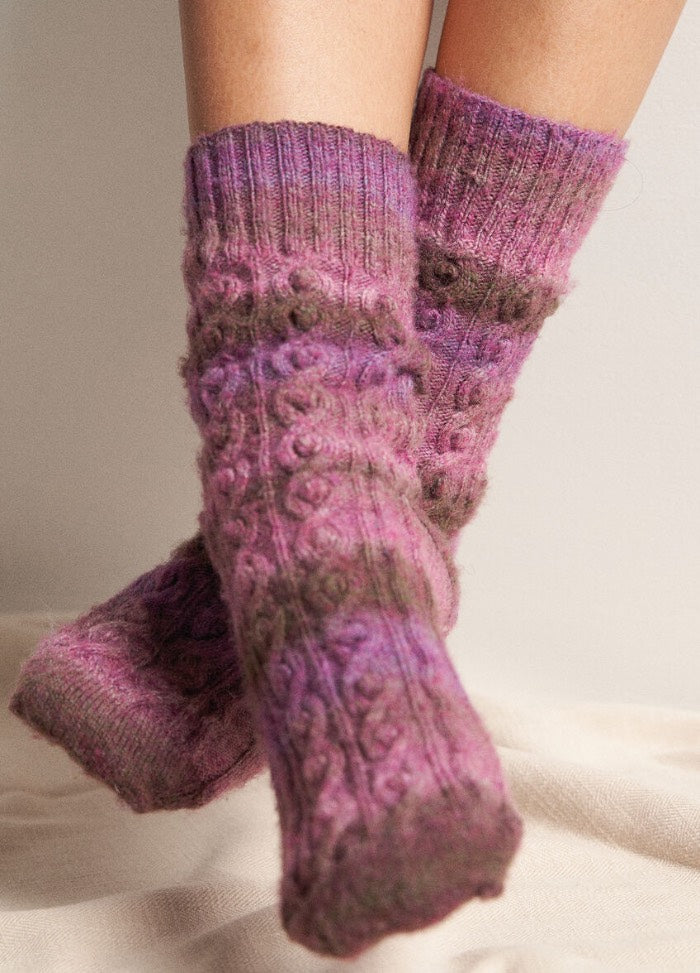 Close-up of legs wearing cozy, knitted socks in shades of purple and brown. The socks feature a cable knit pattern, conveying warmth and comfort.