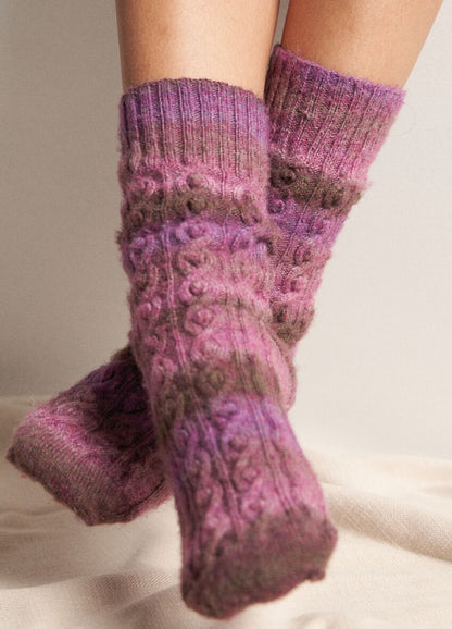 Close-up of legs wearing cozy, knitted socks in shades of purple and brown. The socks feature a cable knit pattern, conveying warmth and comfort.