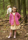 A young girl smiles in a forest, wearing a pink knit cardigan, beige dress, and pink boots. A boy explores the background. Autumn leaves cover the ground.
