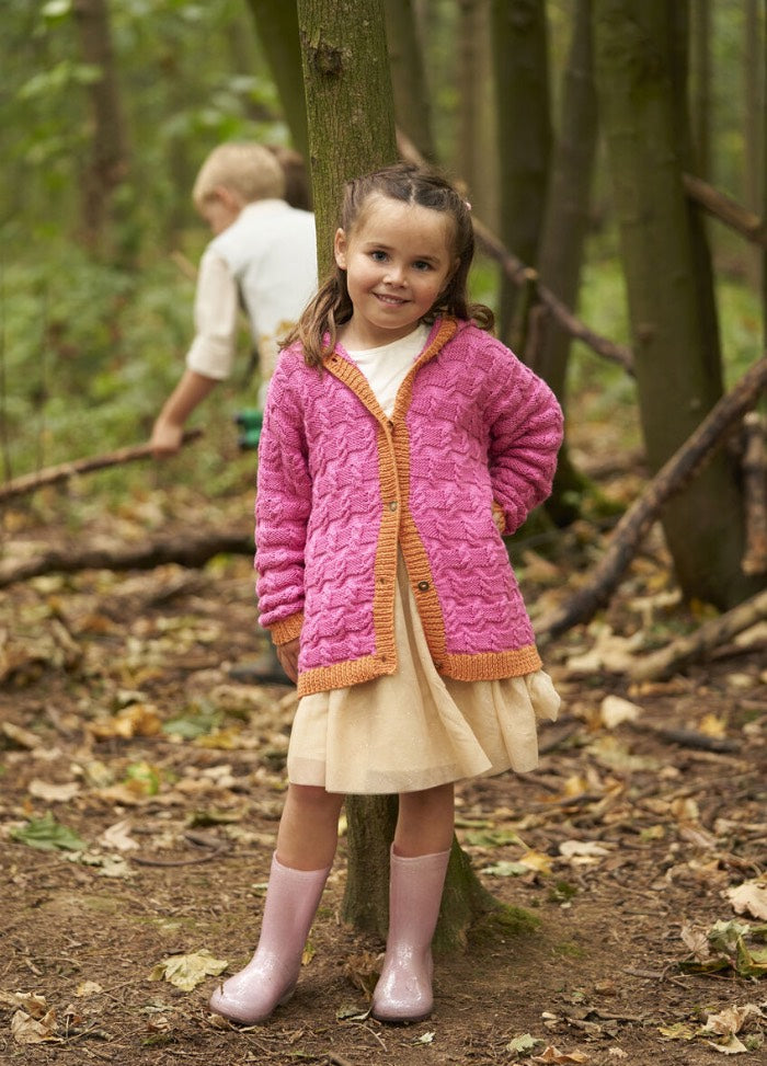 A young girl smiles in a forest, wearing a pink knit cardigan, beige dress, and pink boots. A boy explores the background. Autumn leaves cover the ground.
