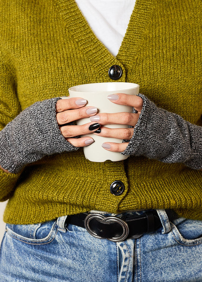 Person wearing gray hand-knitted fingerless gloves, styled with a mustard yellow cardigan and blue jeans, holding a white cup – cozy and functional knitwear for everyday use.